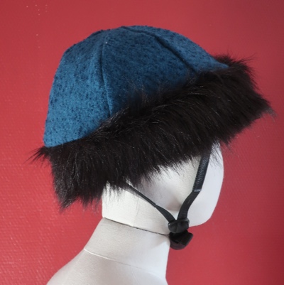 Kuvassa villakankaasta tehty turkoosi kypäränpäällinen, jossa on musta karvareunus.